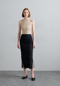 Beige aansluitend tanktop met tekst, gecombineerd met een zwarte knie-lange rok met zijslits. Model staat op een grijze vloer tegen een lichte muur.