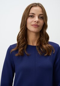 Top blu navy con scollatura tonda e maniche lunghe, realizzato in un tessuto liscio. Presenta dettagli di cucitura discreti sul davanti.