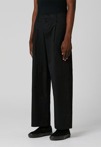 Black wide-leg trousers made of smooth fabric with a neutral finish. Features front pleats and a tailored fit, paired with black shoes.