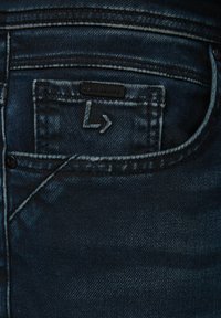 Granatowe dżinsy z widoczną fakturą, z tylną kieszenią, metką z logo i kontrastowym przeszyciem. Materiał wydaje się lekko zużyty.