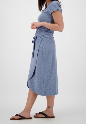 Vrouw die een blauw shirt met korte mouwen en bijpassende wikkelrok met strikceintuur draagt, zijwaarts staand met zwarte sandalen op een witte achtergrond.