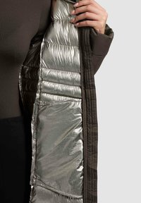 Chaqueta de tela marrón con un forro interior plateado metálico, que presenta secciones acolchadas y un bolsillo escondido en el interior.