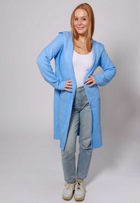 Cardigan con cappuccio di colore azzurro chiaro, realizzato in tessuto morbido, dotato di due tasche frontali, design aperto davanti, abbinato a jeans chiari e sneakers.