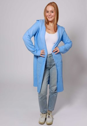 Hellblauer Kapuzencardigan aus weichem Material, mit zwei vorderen Taschen, offenem Frontdesign und kombiniert mit hellen Jeans und Sneakern.