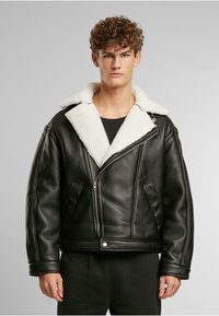 Jongen met krullend haar, gekleed in een zwart leren jack met een witte schapenvacht kraag en zwarte broek, staand tegen een effen lichte achtergrond.