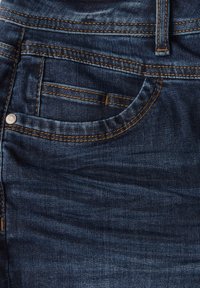 Jeans i mörkblå denim med kontrasterande gul sömnad. Har en framficka och knapp i metall. Texturen är något sliten.