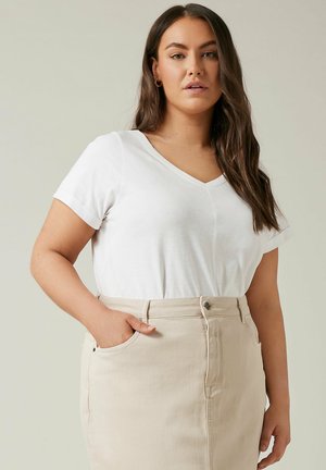 Kobieta o długich brązowych włosach, ubrana w białą koszulkę z dekoltem w serek i beżową spódnicę z wysokim stanem, z jedną ręką w kieszeni, na jednolitym tle.