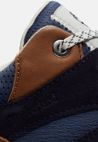 Primer plano de una zapatilla azul y marrón con cordones blancos y negros con patrón, pasados por un ojal metálico.