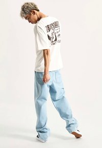 Biały T-shirt z nadrukiem na plecach, jasnoniebieskie luźne jeansy oraz czarno-białe trampki; modelka odwrócona tyłem do kamery.