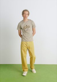 Beige tričko s logem a grafickým designem, žluté široké kalhoty a bílé tenisky. Hladká textura, volný střih a uvolněný styl.