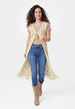Kvinde med krøllet hår går, iført en beige skjorte, blå jeans, beige støvler og en gul hæklevest med frynser.