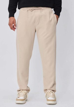 Homme portant un pantalon beige à cordon, des baskets blanches et beiges, et un haut noir à manches longues, debout les mains le long du corps.