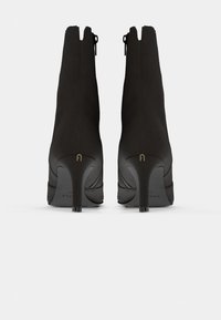 Botas de tornozelo pretas com uma textura de camurça suave, fecho de zip na parte de trás, bico apontado e um salto stiletto moderado. Detalhe do logotipo dourado no salto.