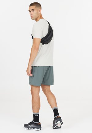 Sort crossover-taske båret over en lys grå skjorte, med grønne shorts og sorte løbesko; har lynlåse og et strømlinet design.