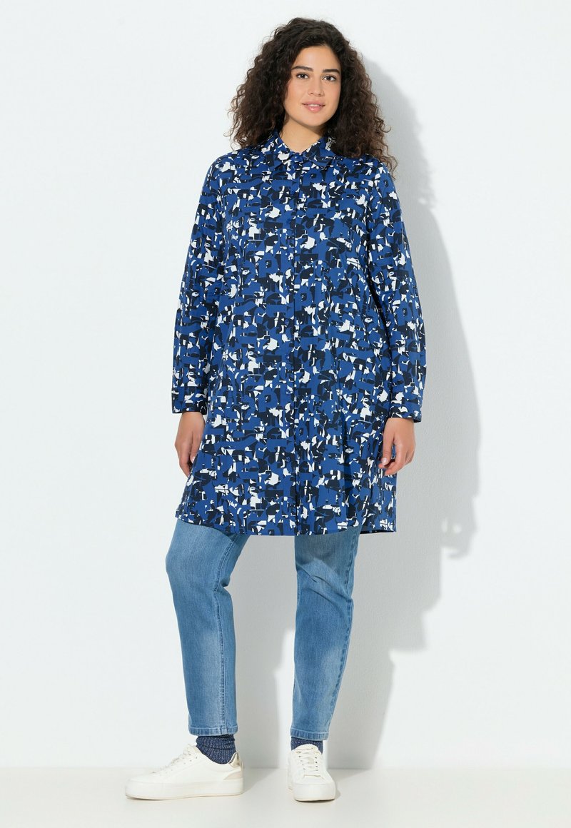 Blauw overhemdjurk met abstracte witte en marineblauwe patronen, voorzien van een kraag, lange mouwen en een losse pasvorm, gecombineerd met lichte spijkerbroek en witte sneakers.