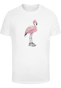 Bílá bavlněná tričko s grafikou růžového plameňáka nosícího tenisky. Design obsahuje podrobné peří a černý zobák.