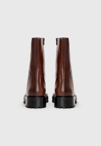 Par črnih usnjenih gležnjarjev s črnimi debelimi podplati in zadrgami na hrbtni strani, prikazanih od zadaj na belem ozadju.