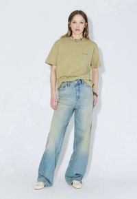 T-shirt manches courtes vert olive avec un petit logo, accompagné d'un jean denim bleu clair ample. Des baskets blanches complètent le look.
