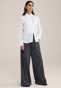 Biela blúzka s dlhým rukávom a golierom, skombinovaná so šedými širokými nohavicami. Blúzka má hladký povrch a detaily s gombíkmi.
