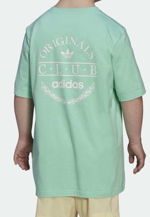 Dziecko noszące miętowo zieloną koszulkę Adidas Originals Club z białym logo i beżowe szorty, widziane od tyłu.