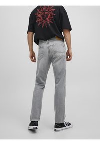 Hombre vistiendo jeans grises claros, camiseta negra con un gráfico de sol rojo en la parte posterior y zapatillas negras, de pie contra un fondo gris liso.