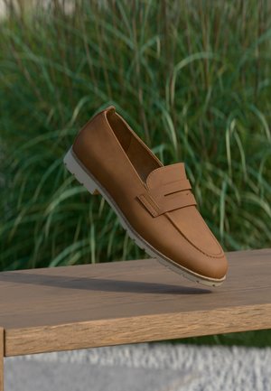 Mocassin en cuir marron avec semelle blanche posé en équilibre sur une surface en bois, hautes herbes vertes en arrière-plan.