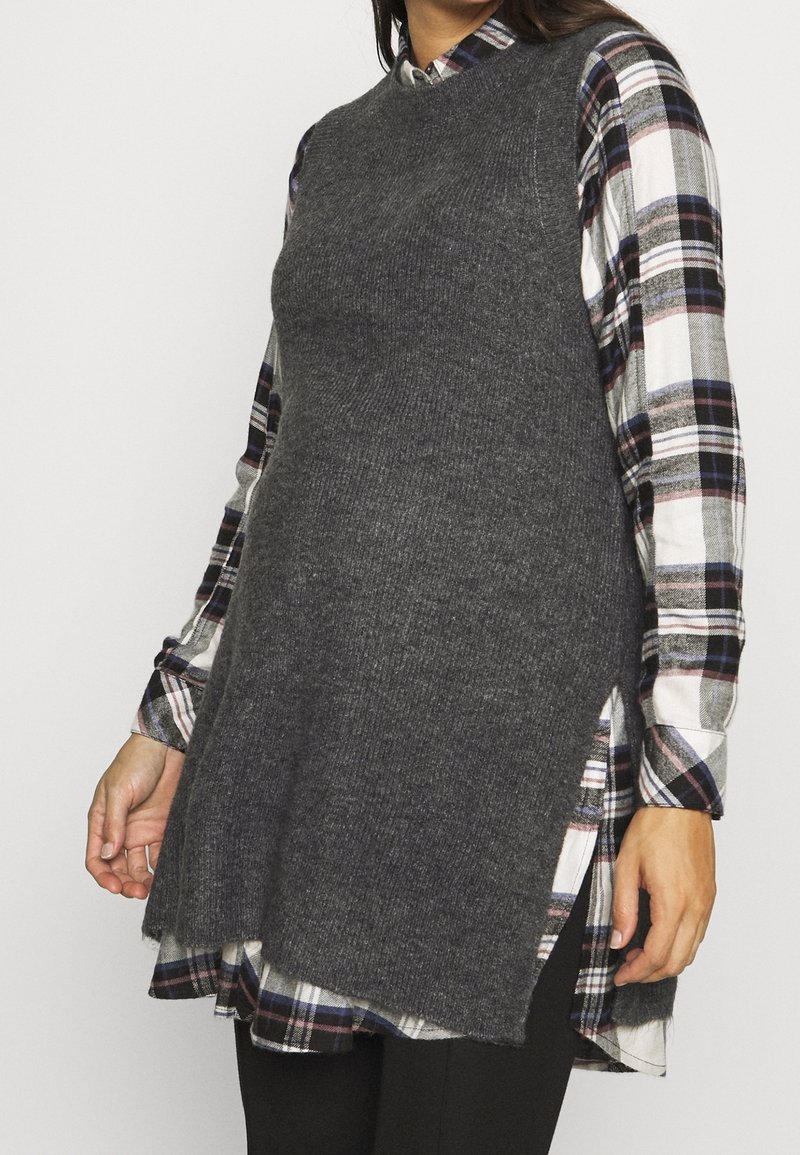 Grijze gebreide trui met een high-low zoom, gedragen over een geruite blouse met blauwe, witte en rode ruitpatronen. Lange mouwen zijn zichtbaar.