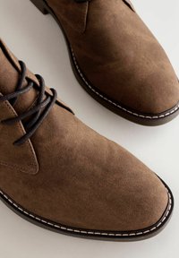 Brązowe zamszowe buty o gładkiej fakturze, okrągłym nosku i kontrastowym przeszyciu na podeszwie. Posiadają ciemne sznurowadła oraz klasyczny design.