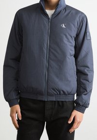 Granatowa pikowana kurtka z wysokim kołnierzem, zamkiem z przodu, elastycznymi mankietami i pasem. Z niewielkim logo na piersi.