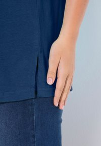 Marineblaue Kurzarmbluse mit seitlichem Schlitzdetail, abgebildet zusammen mit einer Hand, die auf dunklen Jeans mit glatter Textur ruht.