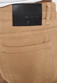 Pantalones de mezclilla color beige con un parche de piel negra en la cintura que muestra las letras "DEF". Presenta bolsillos traseros con costura visible.