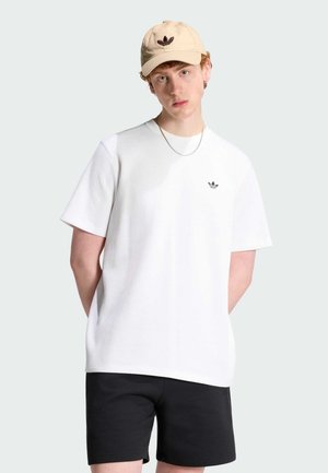 Junger Mann trägt beige Adidas-Kappe, weißes Adidas-ripptes Shirt, schwarze Shorts und silberne Kettenkette, steht mit den Händen hinter dem Rücken.