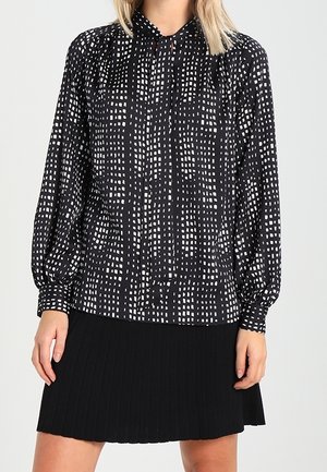 Vrouw draagt een zwarte blouse met witte vierkante patronen en een zwarte geplooide rok tegen een effen achtergrond.