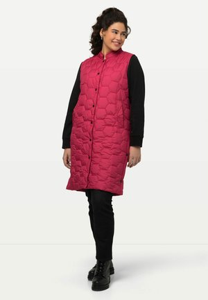 Gilet lungo rosa a piumino con motivo a rombi, bottoni a pressione e colletto alto, indossato sopra una maglietta nera a maniche lunghe e jeans.