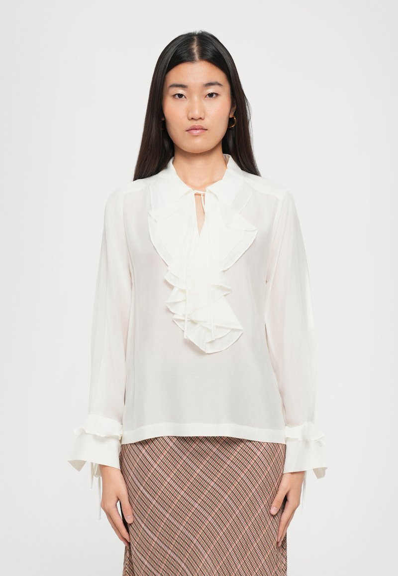 White blouse featuring ruffled front, tie closure at neck, and flared cuffs. The fabric appears lightweight and semi-sheer.