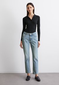 Camisa polo negra de manga larga con canalé, jeans de tiro alto en azul claro y mocasines negros. Los jeans están ligeramente desgastados y tienen un corte recto.