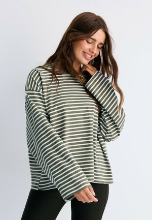 Woman smiling down wearing loose green and white striped long-sleeve top and black pants against a plain light background.