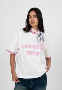 Frau mit langen dunklen Haaren trägt ein weißes T-Shirt mit rosa Kante und dem Schriftzug "PROHIBITED BERLIN", einen blau gemusterten Schal und blaue Jeans, posiert selbstbewusst.