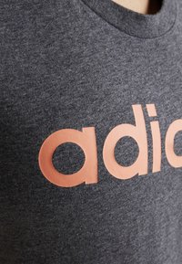 Szara bawełniana koszulka z dużym koralowym logo "adidas". Tekstura jest gładka, a dekolt okrągły.