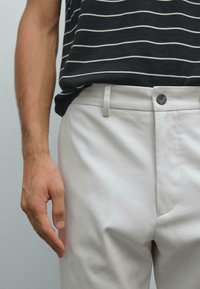 Uomo che indossa pantaloni beige con bottoni e passanti per cintura, e una maglietta nera con sottili righe orizzontali bianche, in piedi contro uno sfondo neutro.