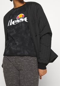 Persona vestida con una chaqueta acolchada negra, una camiseta de tie-dye negra de ellesse con logo y pantalones grises texturizados, de pie contra un fondo simple.