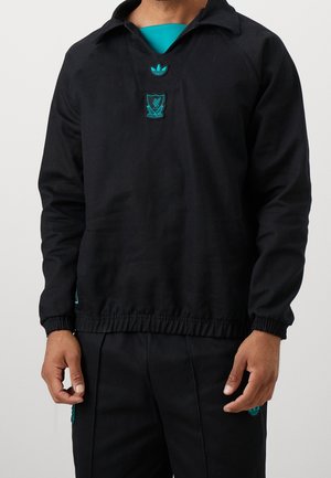 Mann trägt schwarzen Langarm-Pullover mit türkisfarbenen Liverpool FC- und Adidas-Logos, kombiniert mit passenden schwarzen Hosen mit türkisfarbenen Logo-Patches.