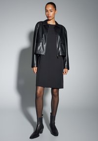 Zwarte leren cropped jas over een zwarte jurk, gecombineerd met zwarte enkellaarsjes. Voorzien van een kraag, zakken en een strak design.