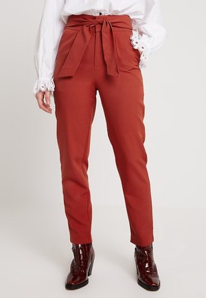 Vrouw draagt roestkleurige broek met hoge taille en gestrikte riem, witte blouse met kanten manchetten en donkerbruine lak enkellaarsjes.
