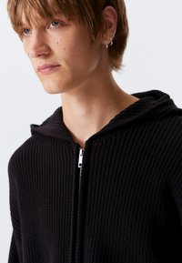 Fekete kötött zipzáras kapucnis pulóver bordázott mintával, kapucnival, magas gallérral és ezüst cipzárral. Egyszerű kialakítás minták vagy díszítések nélkül.