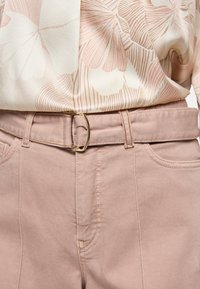 Hellrosa Baumwollhose mit glatter Textur, ausgestattet mit einem Gürtel mit goldener Schnalle und Vordertaschen. Darüber ist eine Bluse mit floralem Muster zu sehen.