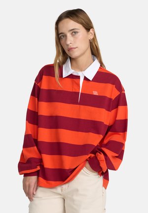 Junge Frau mit langen blonden Haaren, die ein rot-orange gestreiftes Rugby-Shirt und hellbeige Hose vor einem einfarbigen Hintergrund trägt.
