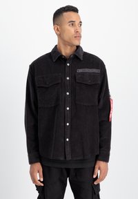 Camicia in cotone corduroy nera con chiusura a bottoni, due tasche sul petto, una toppa con logo e un'etichetta rossa sul maniche sinistro.