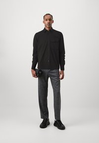 Black long-sleeve shirt with a chest pocket, paired with light gray patterned trousers and black sneakers. Carrying a small black bag.