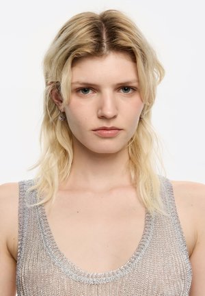 Donna bionda con capelli lunghi fino alle spalle che indossa orecchini a cerchio d'argento e un top senza maniche in maglia metallica su sfondo bianco.